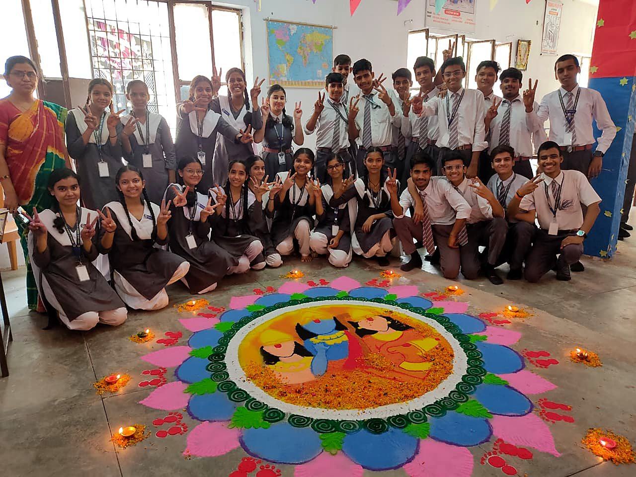 A teacher with smiling senior students in uniform around a colourful rangoli they made in their classroom